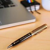 Custom Gold and Black Name Pen with 'Isha Malhotra' engraving, ideal personalized gift, shown on a light wood desk beside a laptop and notebooks.